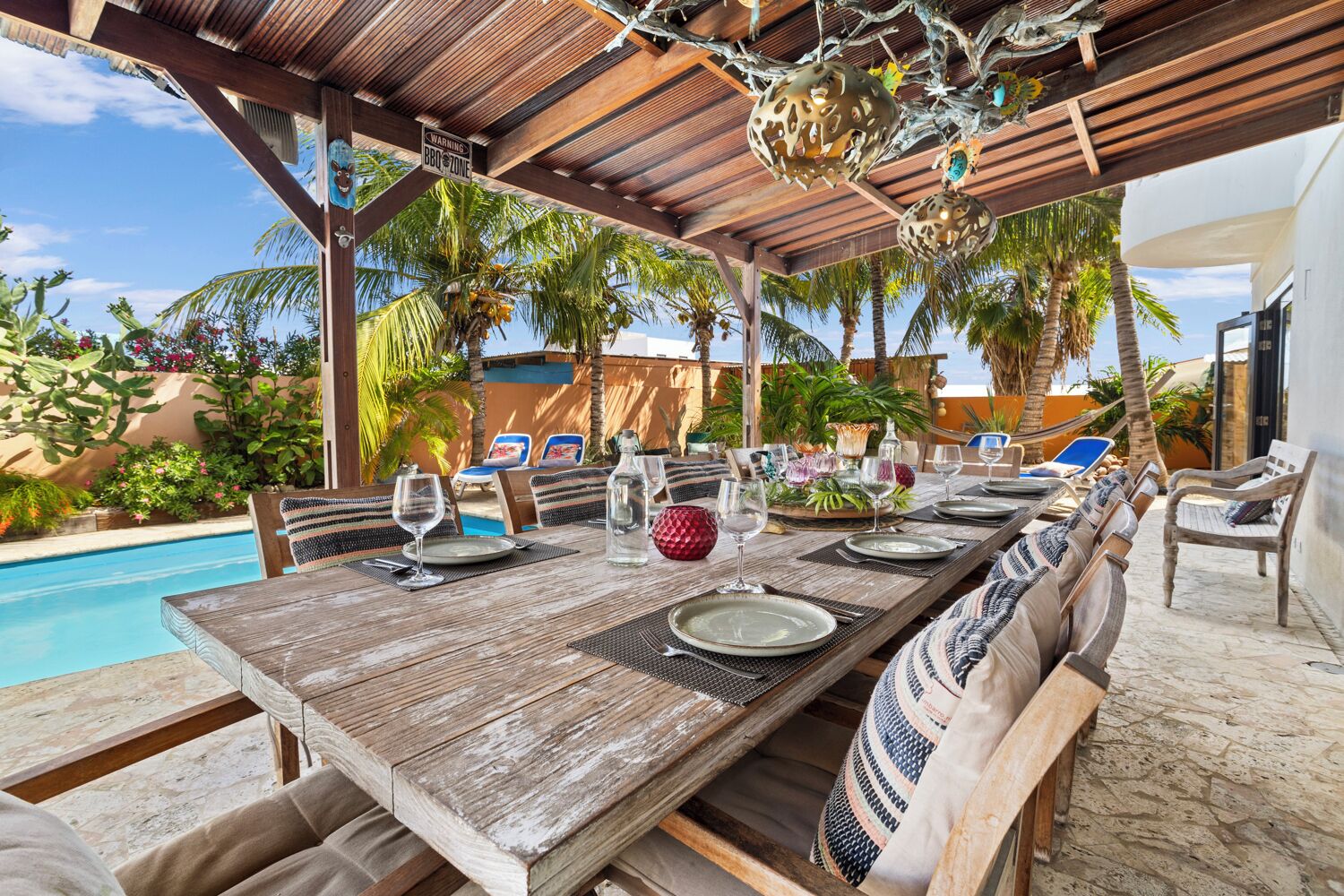 Un espace de restauration en plein air avec une table en bois prévue pour quatre personnes, entourée de chaises en osier, sous une pergola en bois agrémentée d’éléments décoratifs.