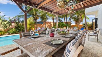 An outdoor dining area with a wooden table set for four, surrounded by wicker chairs, under a wooden pergola with decorative elements.