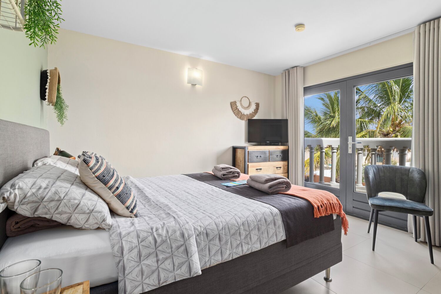 A modern hotel room with a bed, a TV, a balcony with a chair, and a view of palm trees.