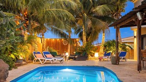 Zona de piscina con sillones reclinables, palmeras y una pérgola de madera.