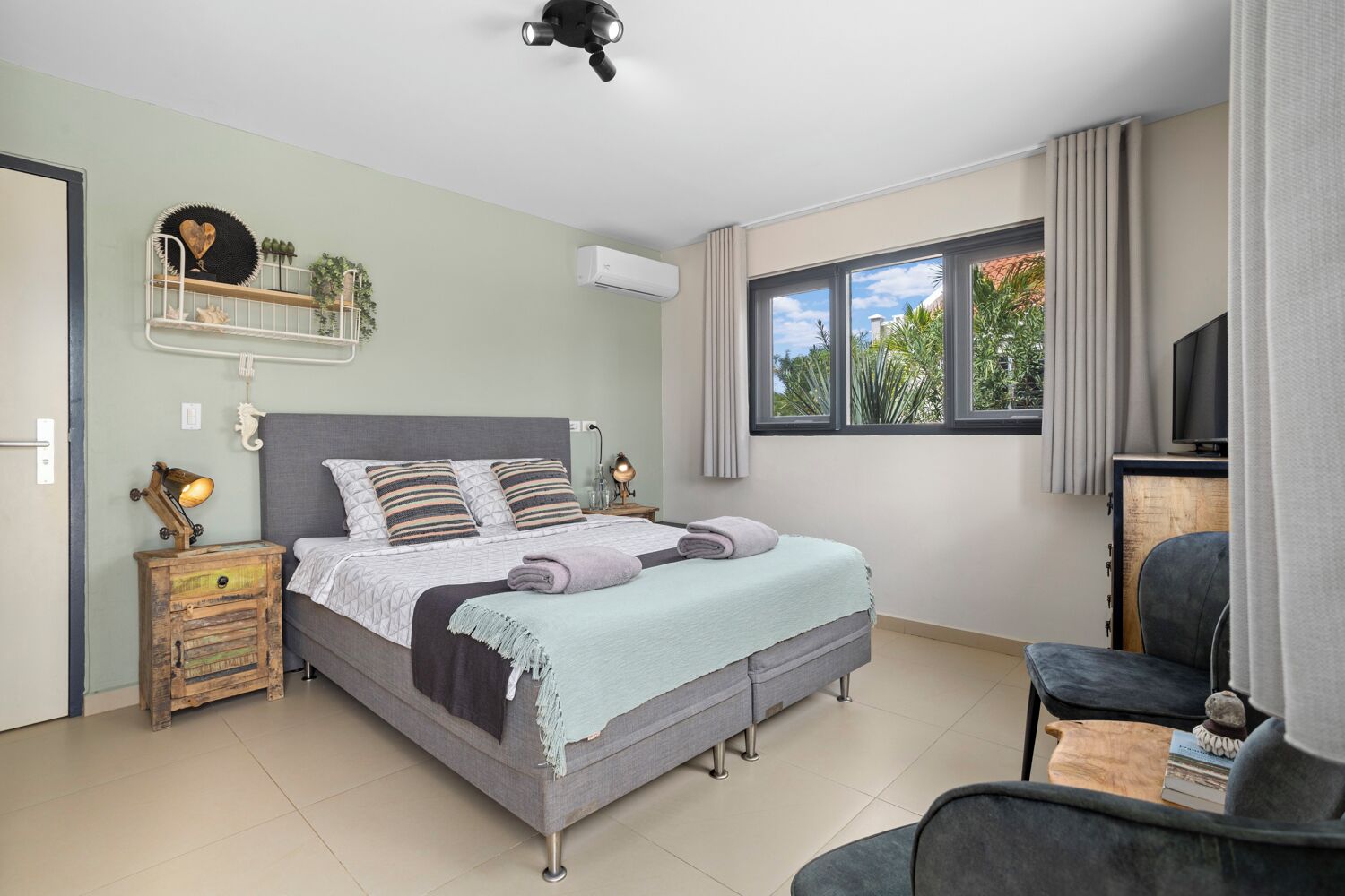 A bedroom with a bed, bedside tables, a chair, a TV, and a window with a view of greenery.