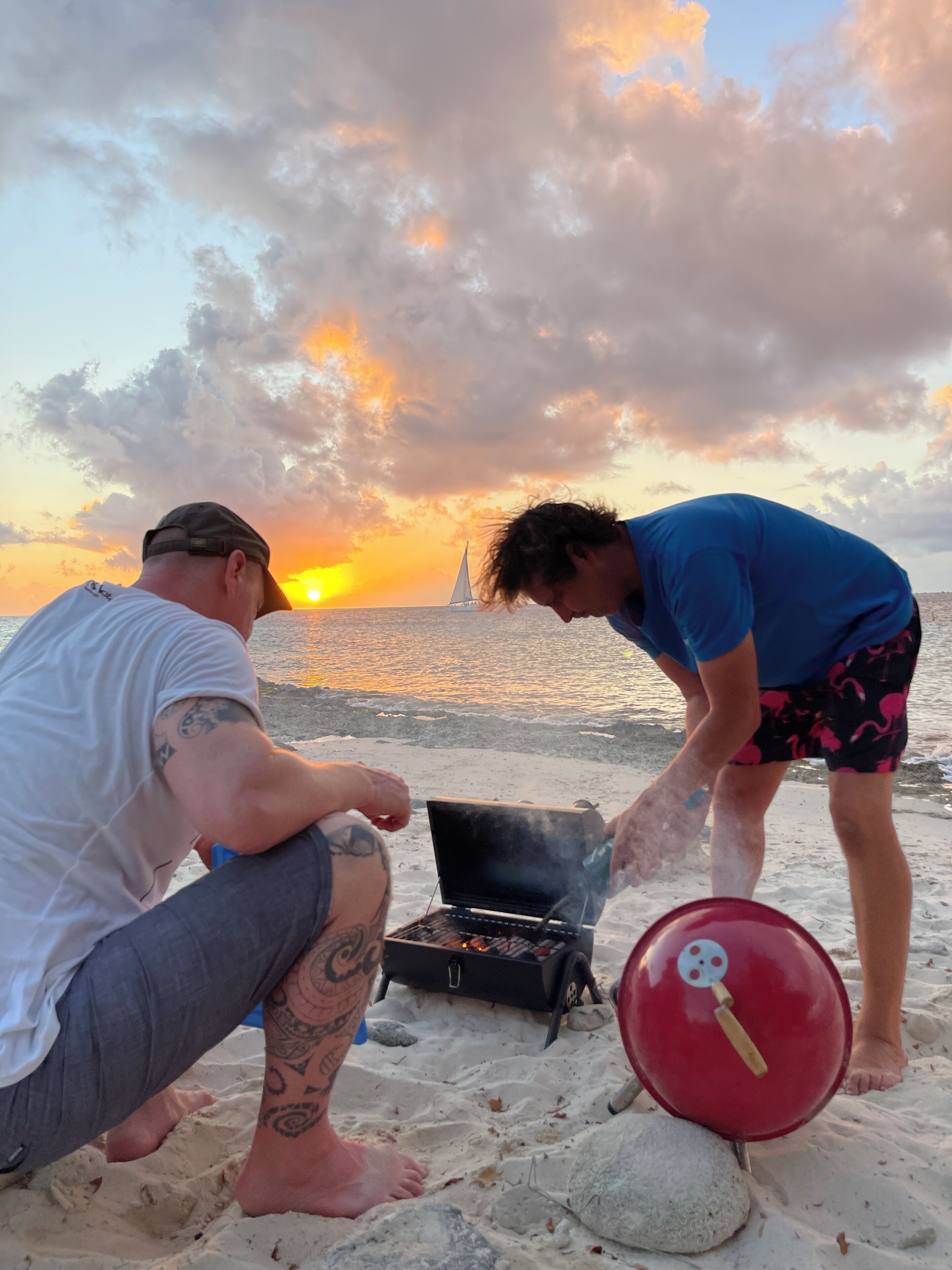 Dos personas cocinando en la playa, con una puesta de sol al fondo.