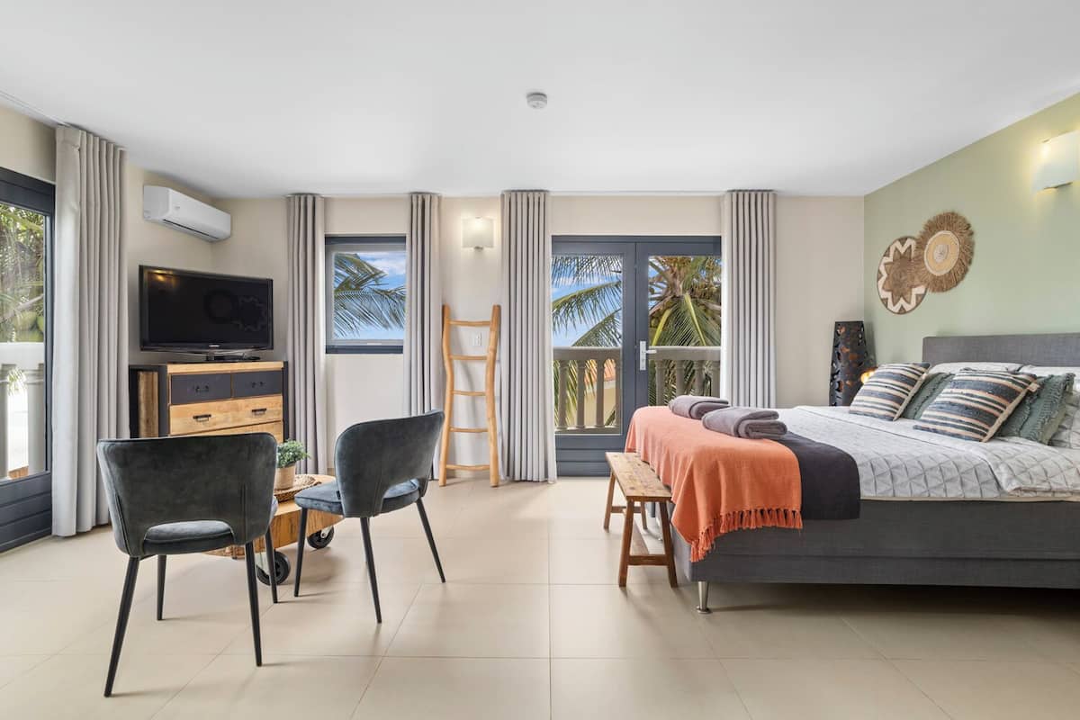 A modern hotel room with a bed, TV, desk, chairs, and a balcony with a view.