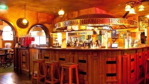Un espace bar avec des tabourets de bar en bois, un comptoir sur lequel est affiché un panneau indiquant « SHENANIGANS », et une décoration intérieure rustique.