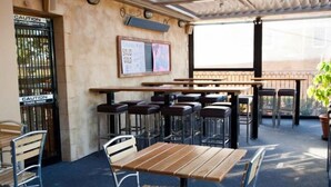 Un café en plein air avec des tables en bois et des chaises en métal, un menu écrit à la craie et un panneau d’avertissement sur le portail.