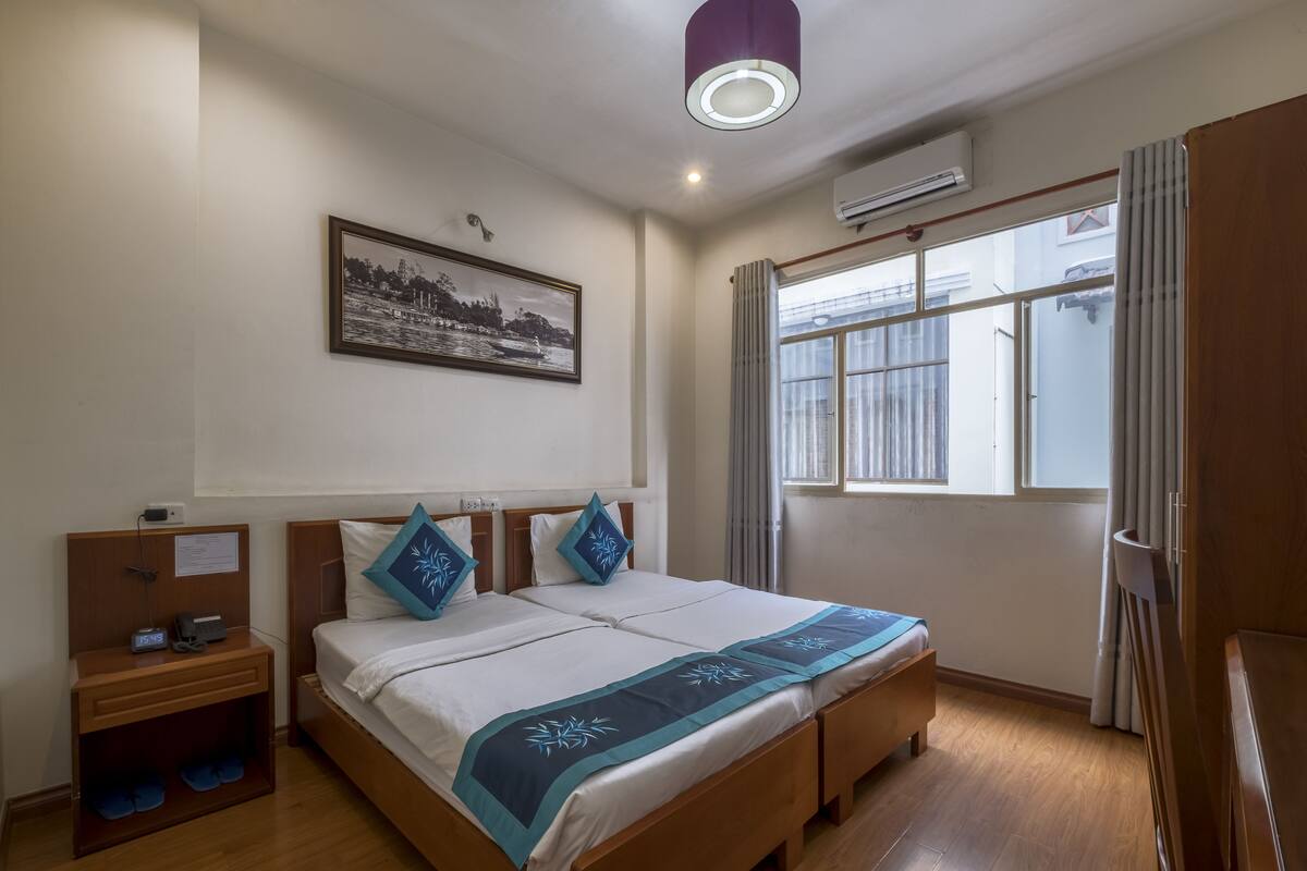 Ein Hotelzimmer mit einem hölzernen Bett, einem Nachttisch, einer Wand-Klimaanlage, einem Fenster mit Vorhängen und einer Deckenleuchte.