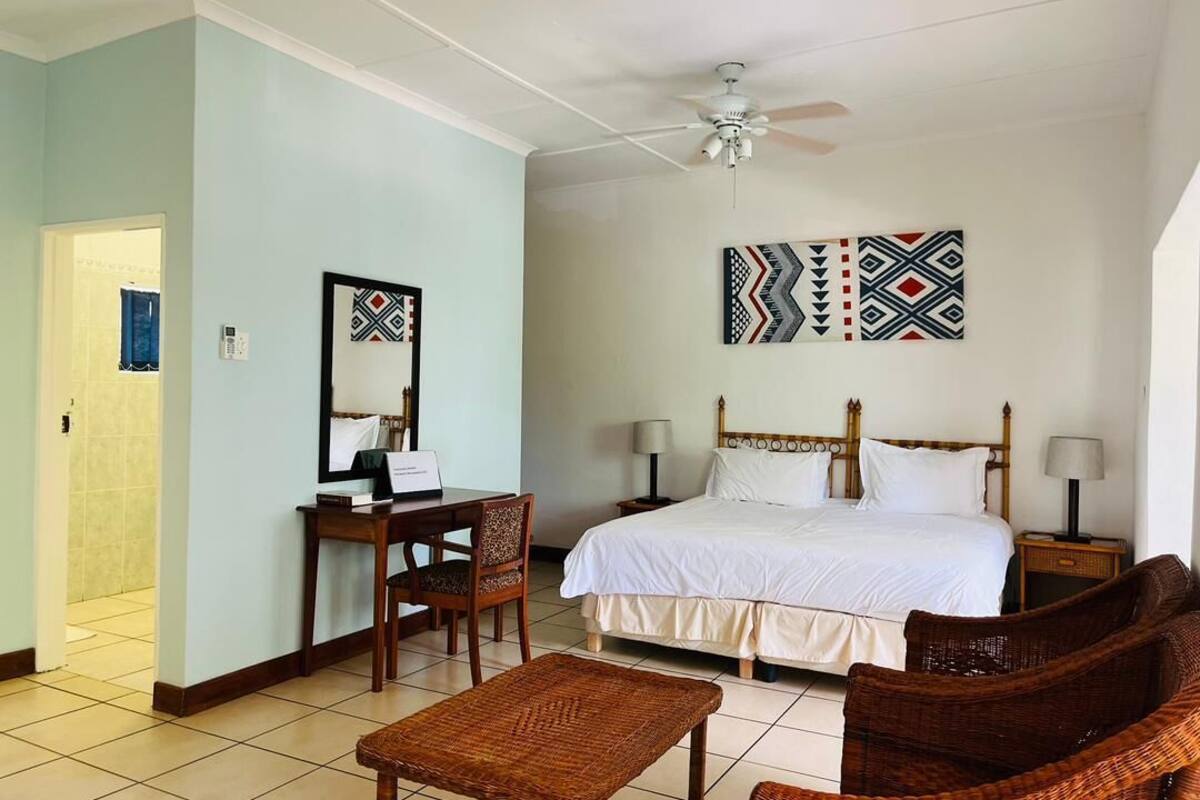 Une chambre d’hôtel comprenant un lit, un bureau avec une chaise, une petite table, deux tables de chevet avec des lampes et un ventilateur de plafond.