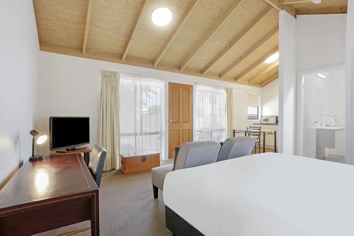 Ein Hotelzimmer mit Holzboden, einem Bett, einem Schreibtisch mit Lampe, einem Fernseher, einem Essbereich mit Stühlen und einer Küchenzeile.