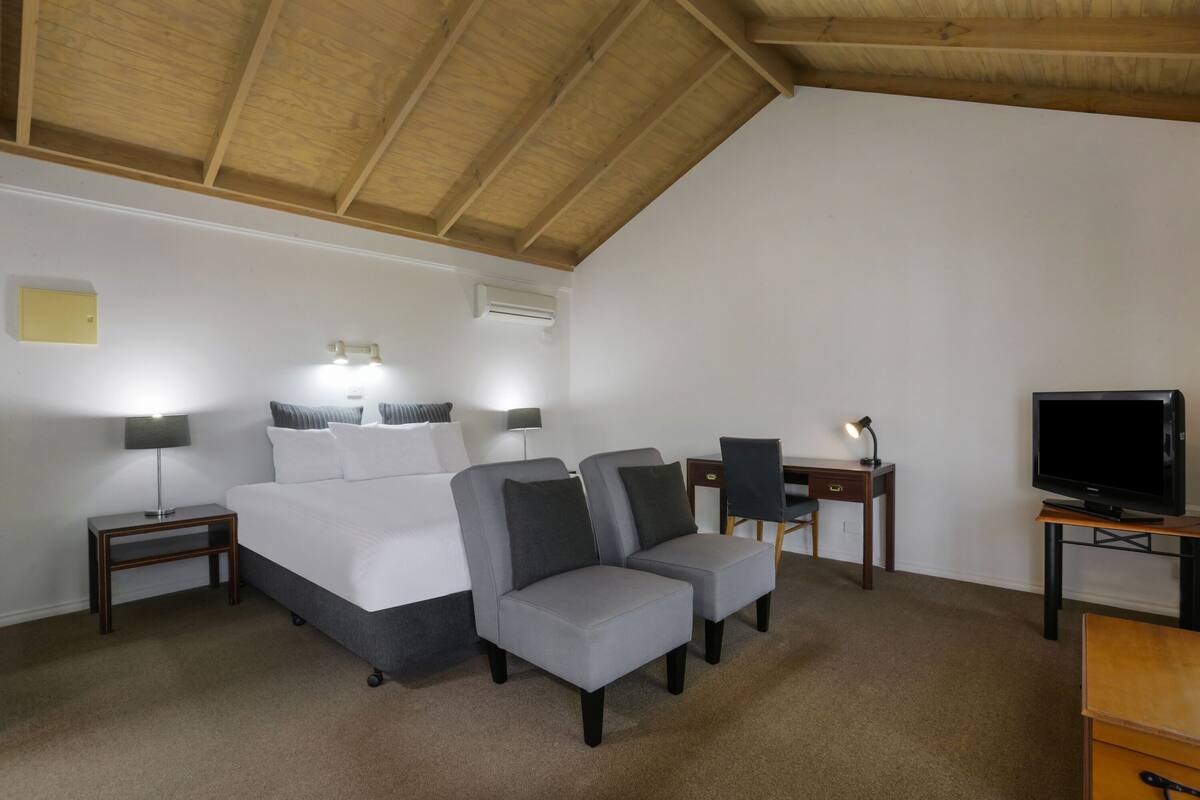 Ein Hotelzimmer mit einem Bett, zwei Sesseln, einem Schreibtisch mit einem Computer, einem Fernseher und einem Deckenventilator.