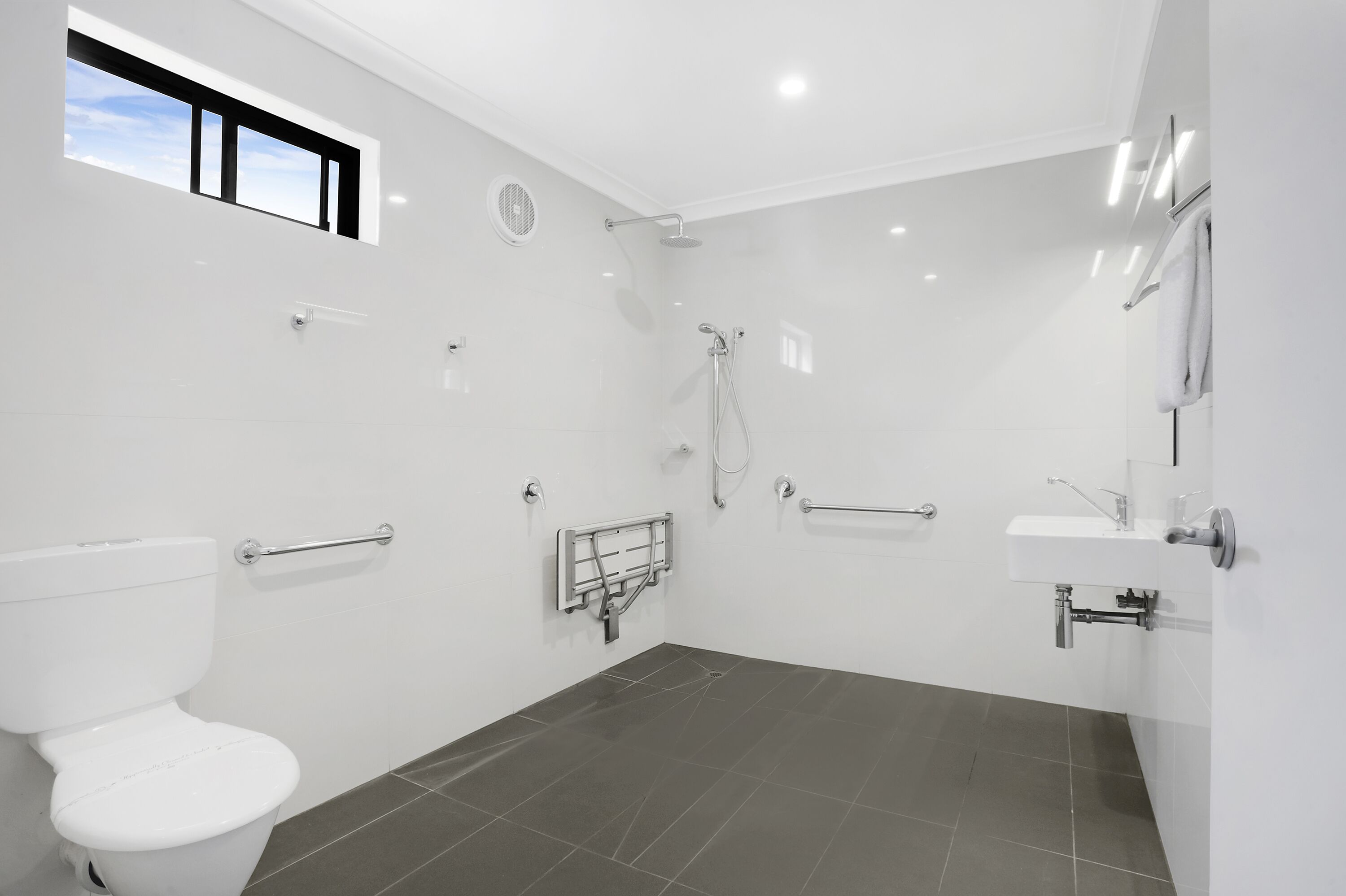 Un baño moderno con inodoro, ducha y lavamanos, equipado con barras de apoyo y una ventana.