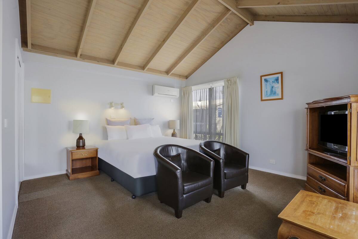 Ein Hotelzimmer mit einem Bett, zwei Stühlen, einem Nachttisch, einer Lampe, einem Fernseher und einem Holzschrank.
