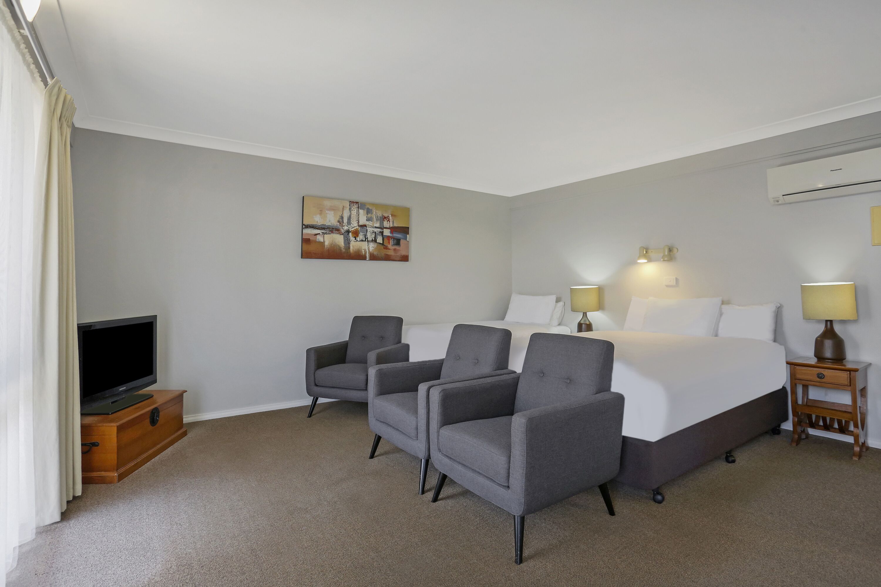 Una habitación de hotel con una cama, dos sillones, un televisor sobre un mueble de madera, un cuadro en la pared y una lámpara en una mesita de noche.