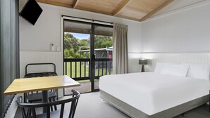 Habitación de hotel con cama, escritorio y silla. Puerta corrediza de vidrio que da a un espacio exterior con vistas a zonas verdes.