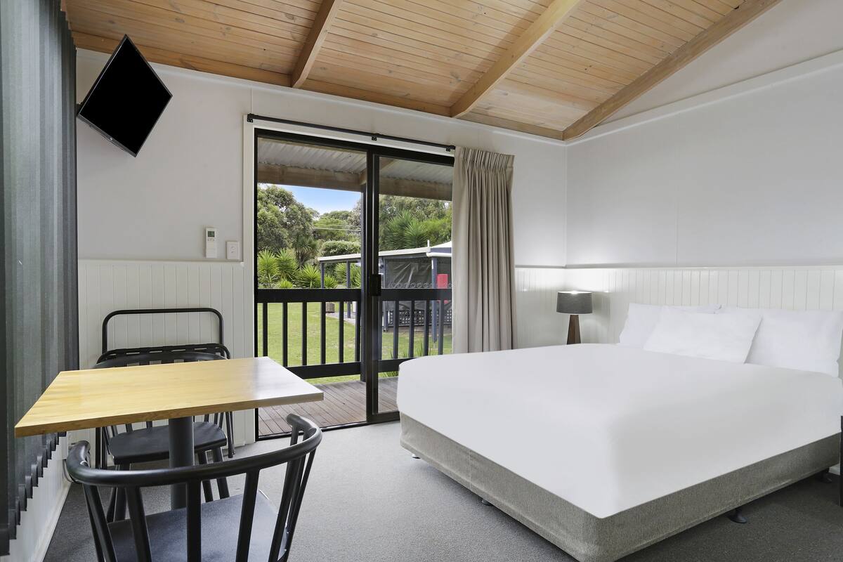Ein Hotelzimmer mit Bett, Schreibtisch und Stuhl. Eine Glastür führt zu einem Außenbereich mit Blick ins Grüne.