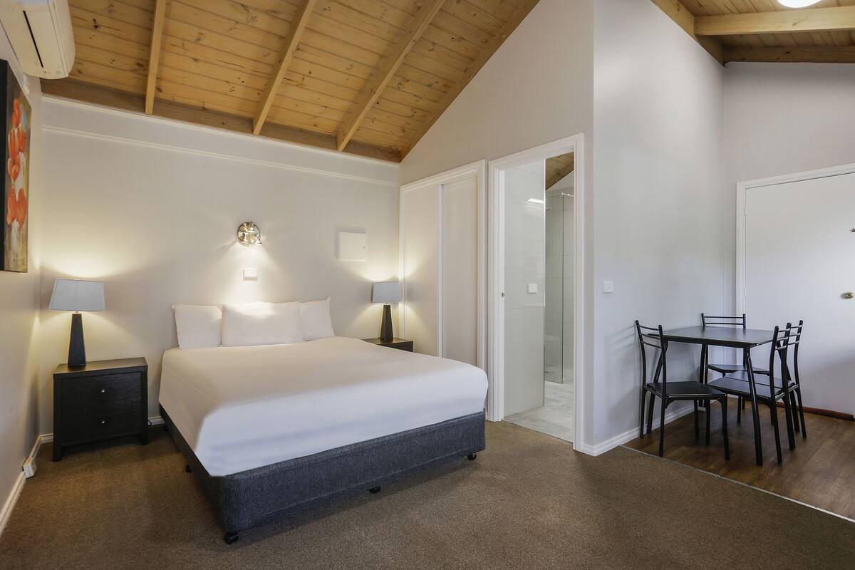 Ein Schlafzimmer mit Bett, Nachttischlampen, einem Nachttisch, einer Holzbalkendecke und einem Essbereich mit Stühlen und Tisch.
