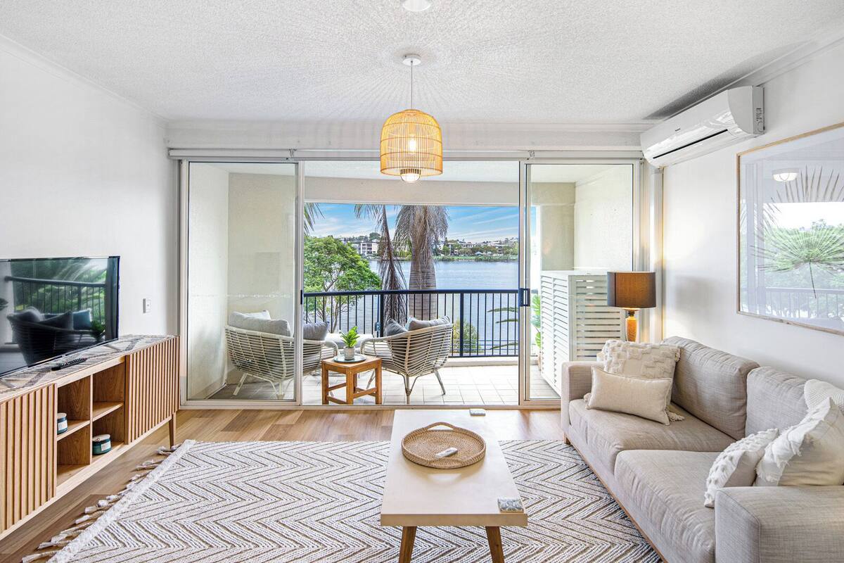Un salon moderne avec un canapé, une table basse et un téléviseur posé sur une étagère en bois. Il y a un balcon avec vue sur l’eau et les arbres.