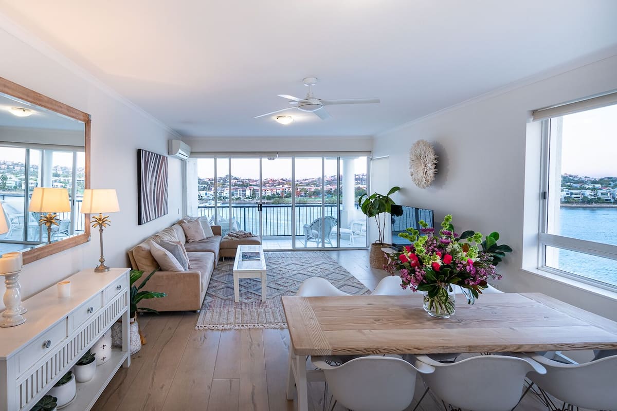 Un salon moderne comprenant un canapé, une table basse, une table à manger et des chaises. Une grande fenêtre offre une vue sur la mer et les bâtiments.