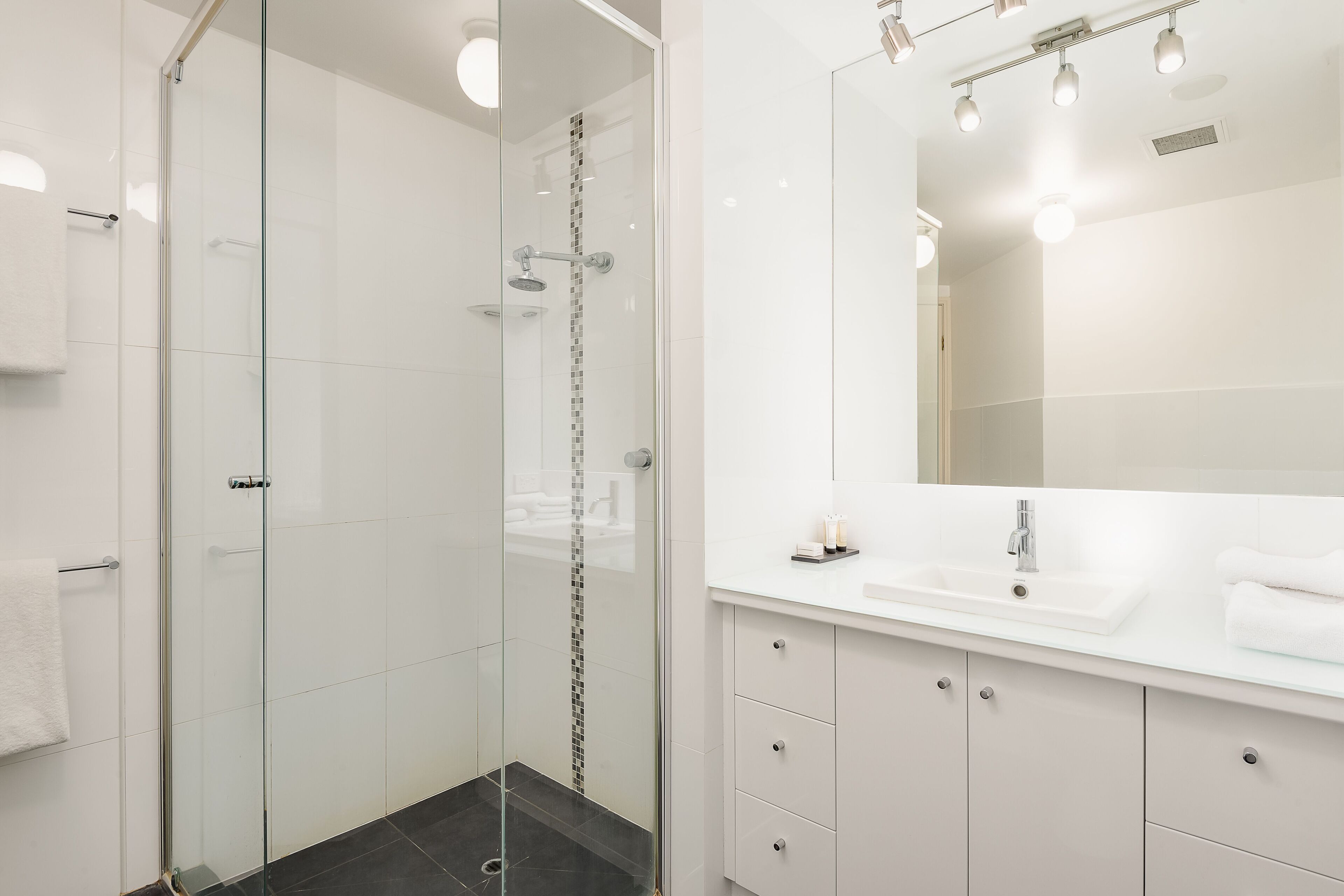 Een moderne badkamer met een douchecabine van glas, een witte wastafel met spiegel erboven en een betegelde vloer.
