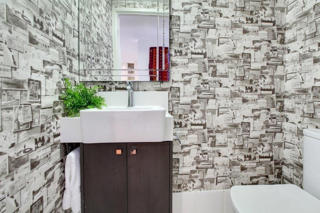 Un baño con lavamanos blanco, mueble de madera oscura y un espejo encima. Las paredes están decoradas con un collage de fotografías en blanco y negro.