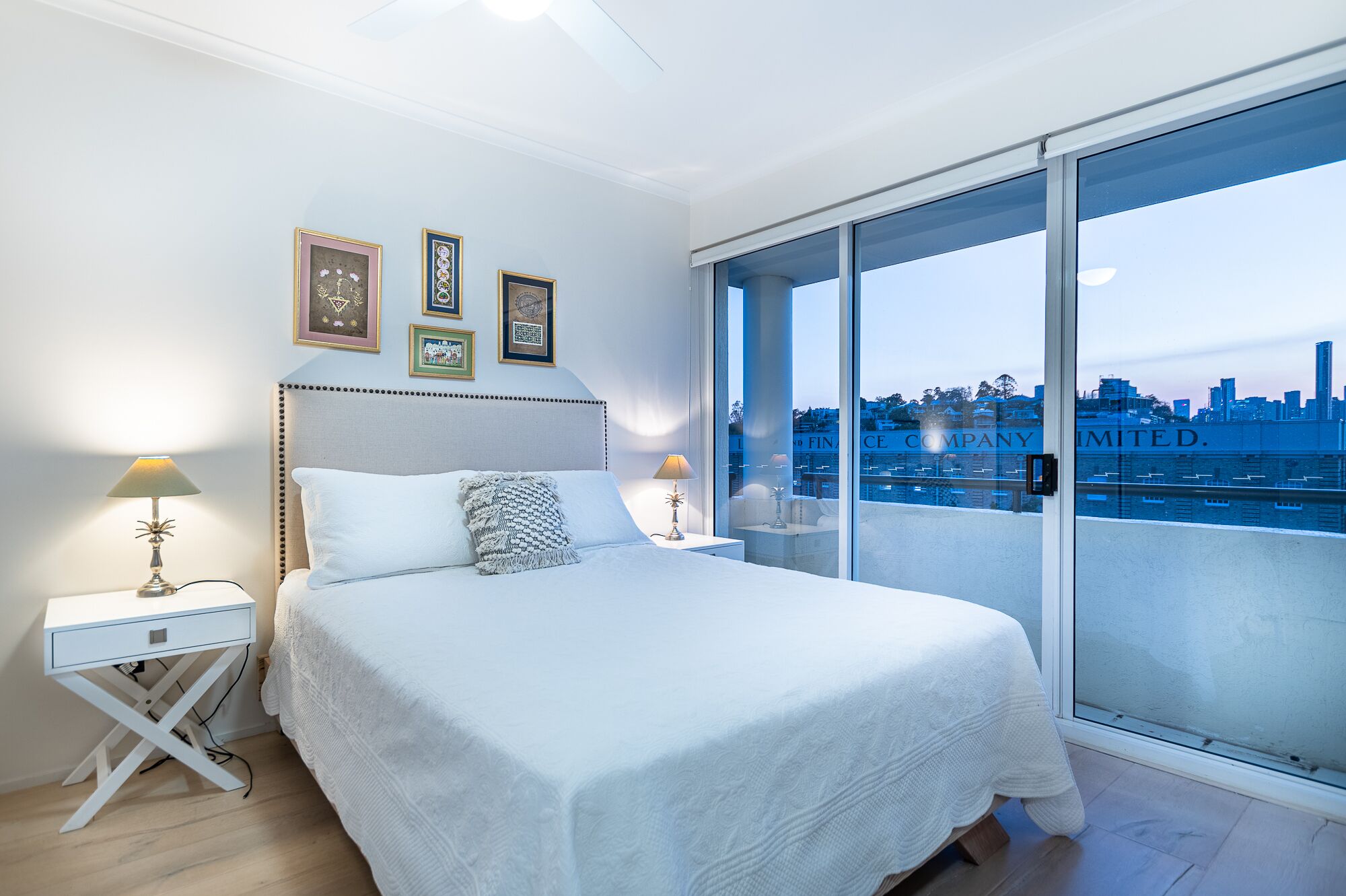 Une chambre moderne avec un grand lit, deux tables de chevet et une vue sur le paysage urbain grâce à de grandes fenêtres du sol au plafond.