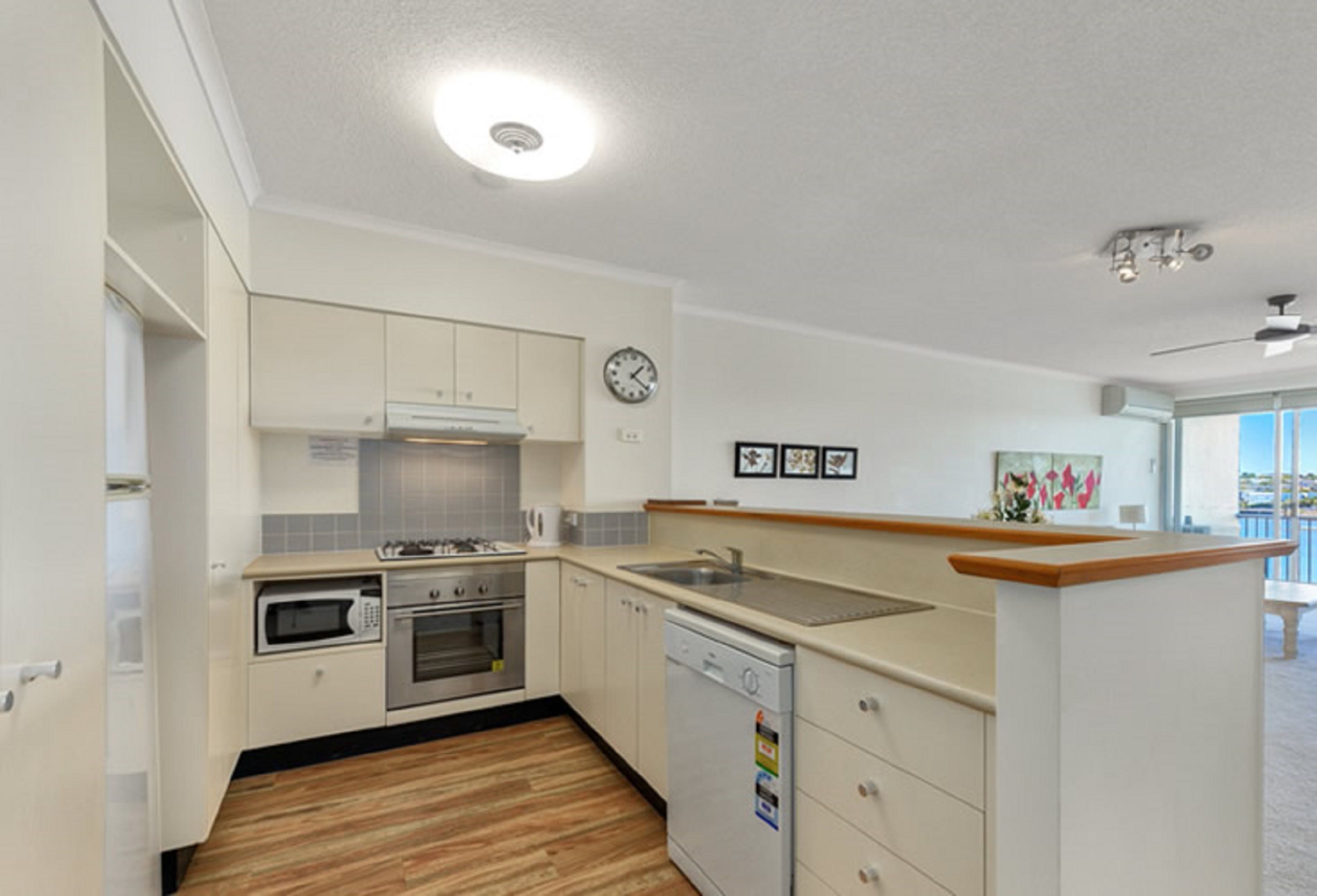 A compact kitchen with white cabinets, a microwave, oven, and dishwasher. There is a clock on the wall and a small window with a view.