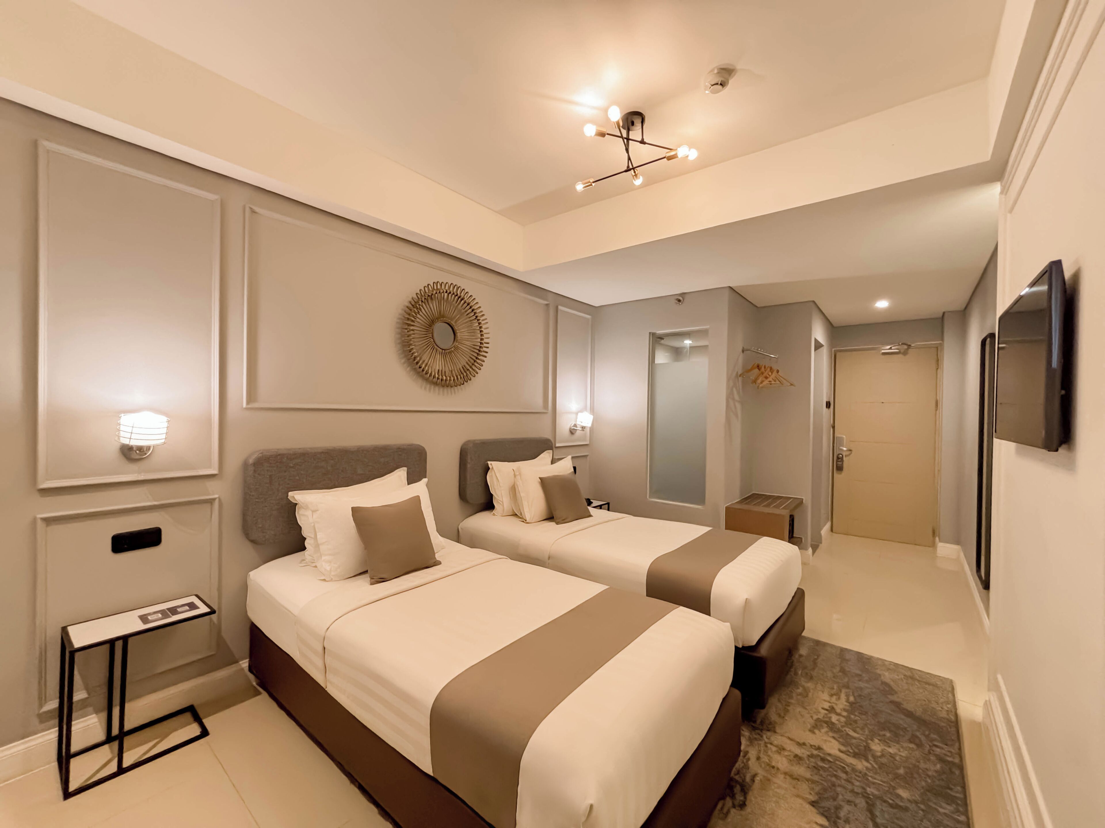 Ein Hotelzimmer mit zwei Betten, einem kleinen Tisch, einem Spiegel, einer Deckenleuchte und einer Tür zu einem weiteren Raum.