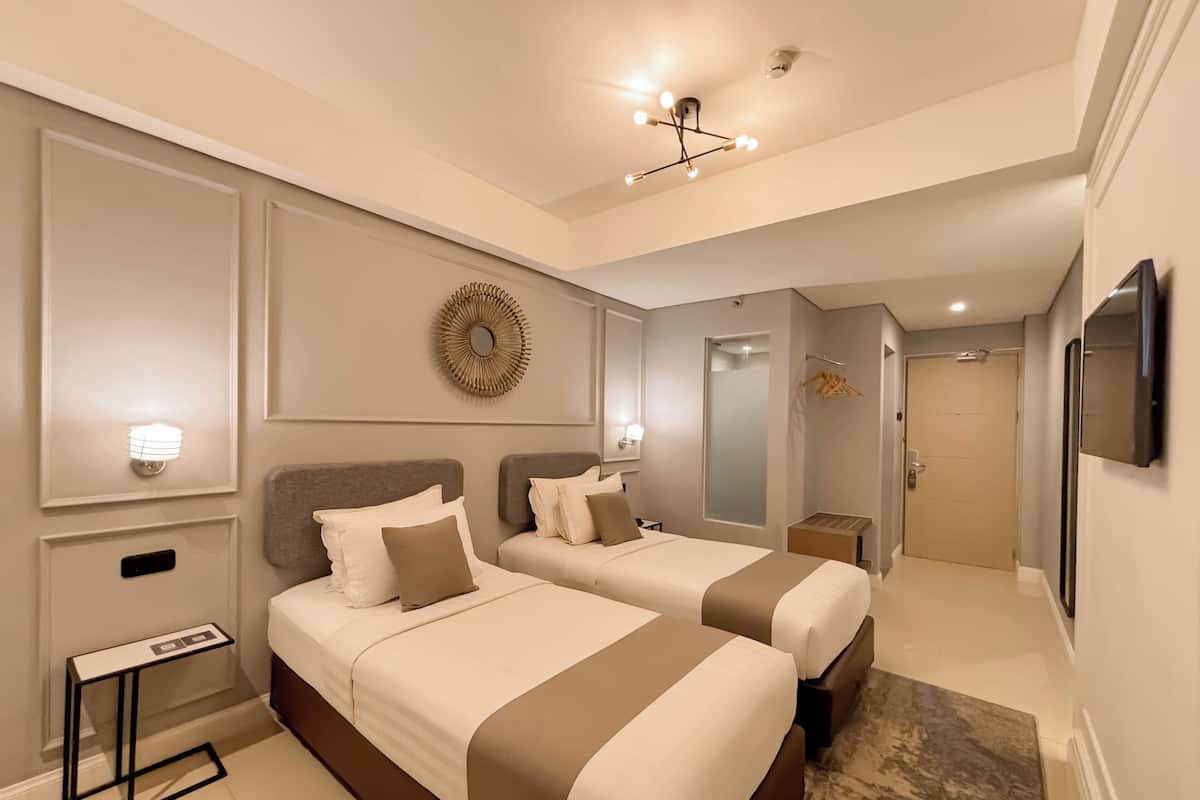 A hotel room with two beds, a small table, a mirror, a ceiling light, and a door leading to another room.
