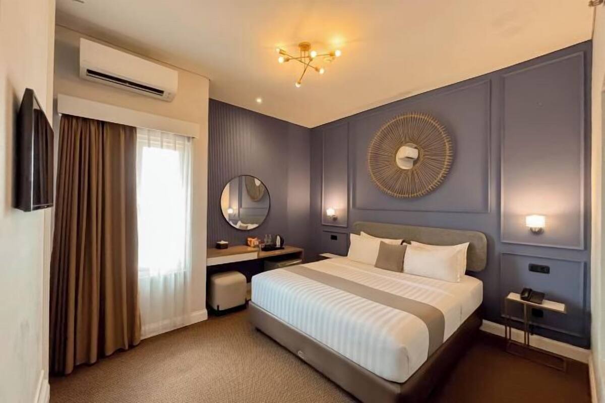A hotel room with a bed, bedside table, mirror, wall-mounted light, and an air conditioning unit.