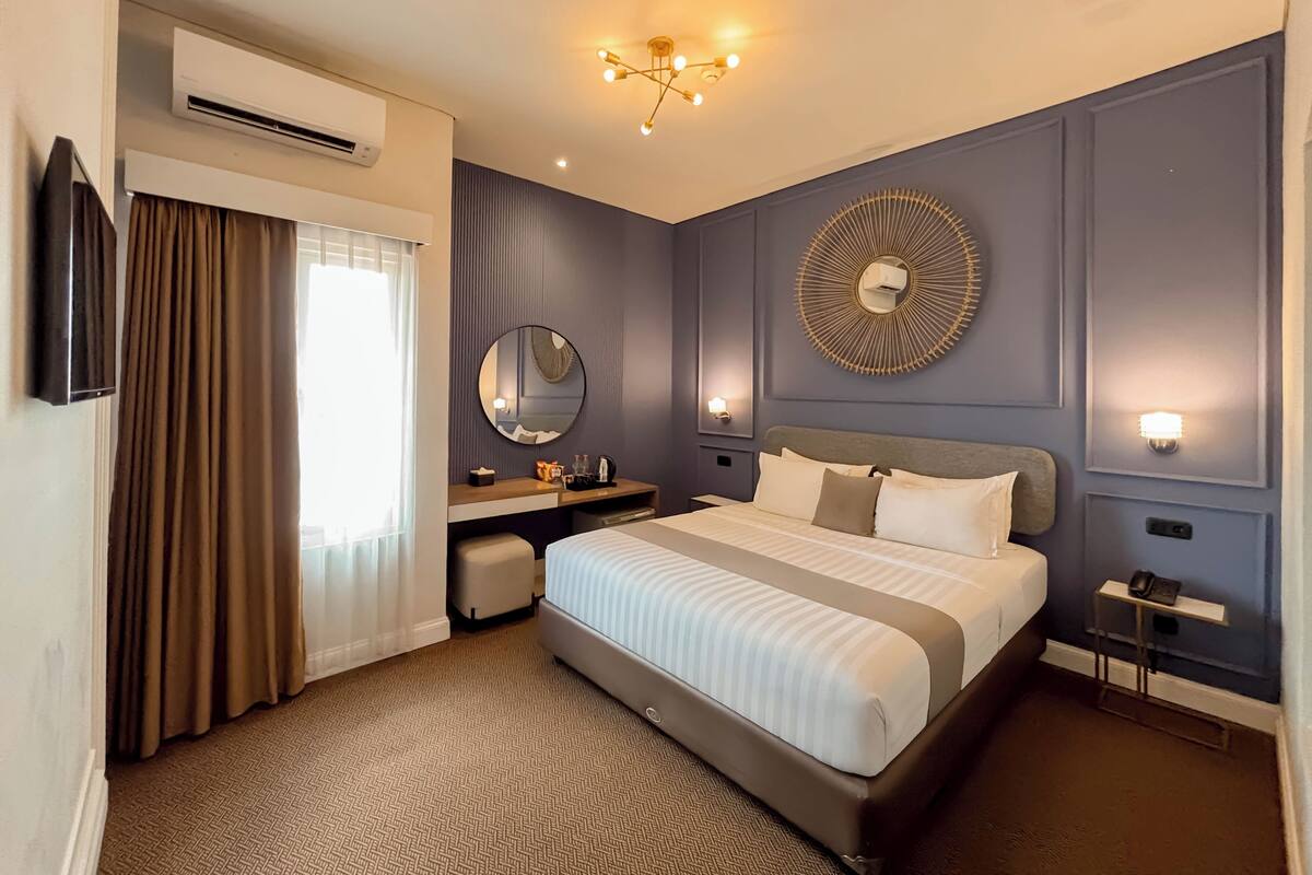 A modern hotel room with a large bed, a desk, a chair, a round mirror, and a wall-mounted light fixture.