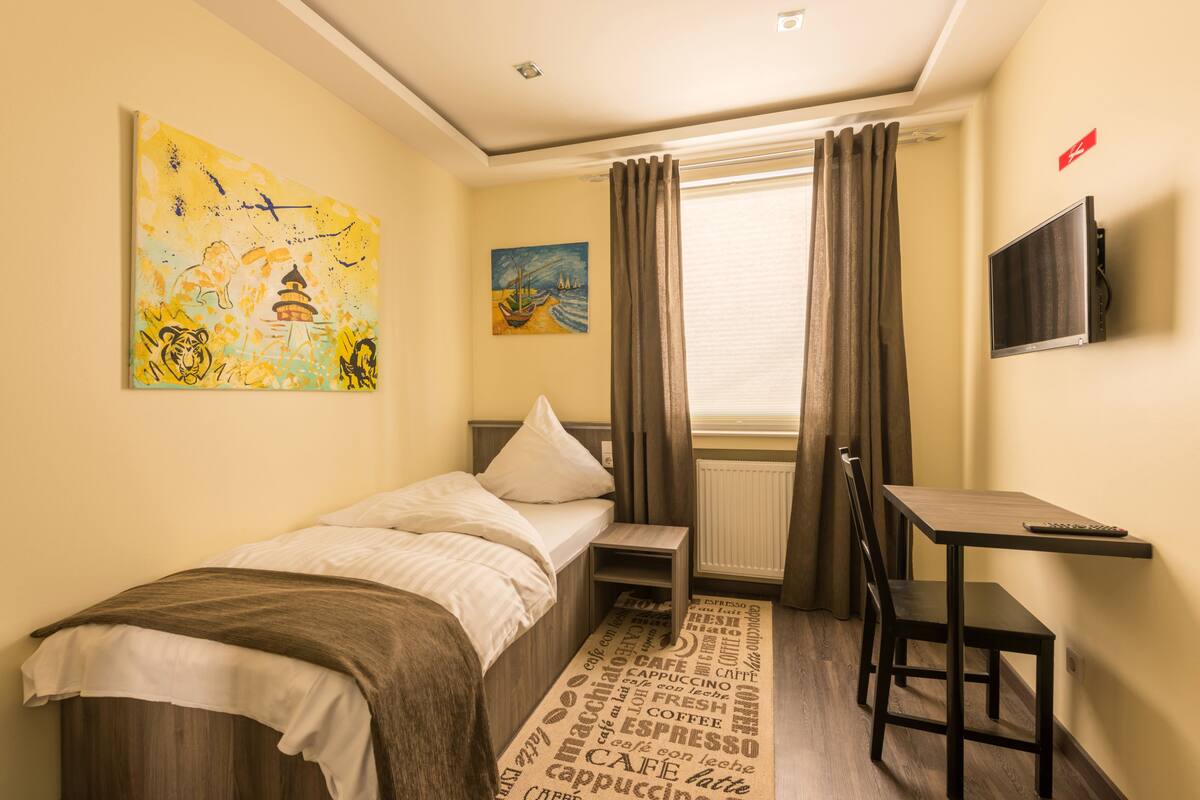 A small, neatly arranged bedroom with a bed, a desk, and a chair. There are two paintings on the wall, a window with curtains, and a television mounted on the wall.