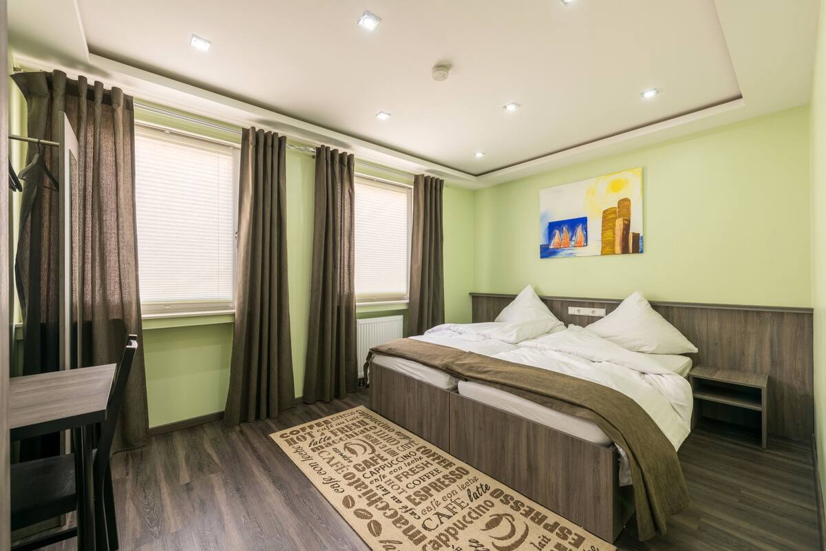 A hotel room with a bed, a bedside table, a chair, a window with curtains, and a painting on the wall.