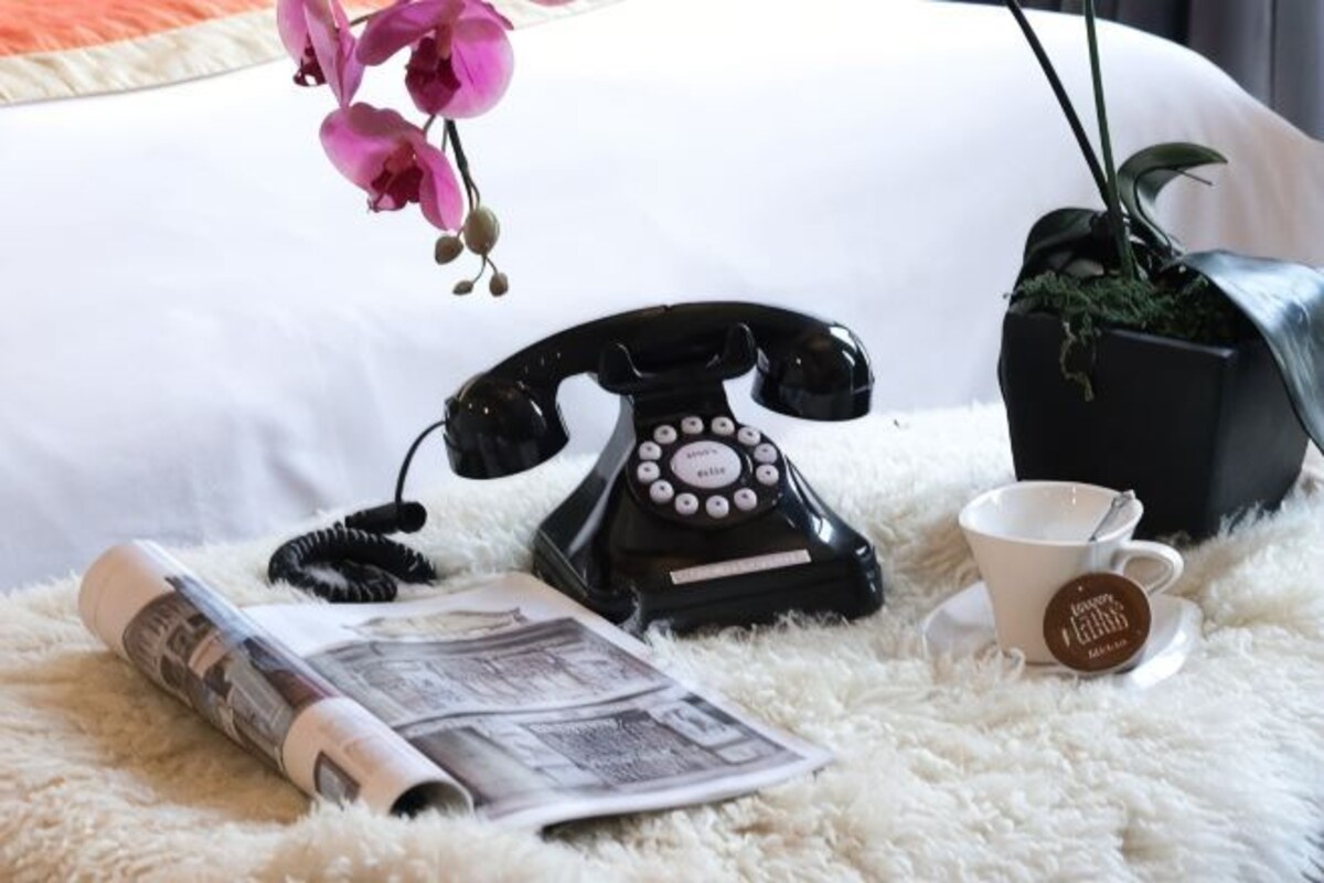 Un teléfono rotativo negro, una taza blanca con plato, un periódico enrollado y una planta en maceta sobre una alfombra blanca mullida.