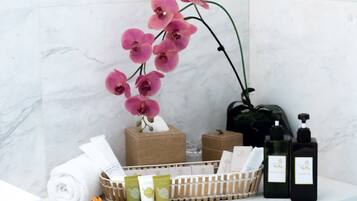 A bathroom with a marble wall, a potted orchid, a towel, a basket of toiletries, and two bottles of shampoo.