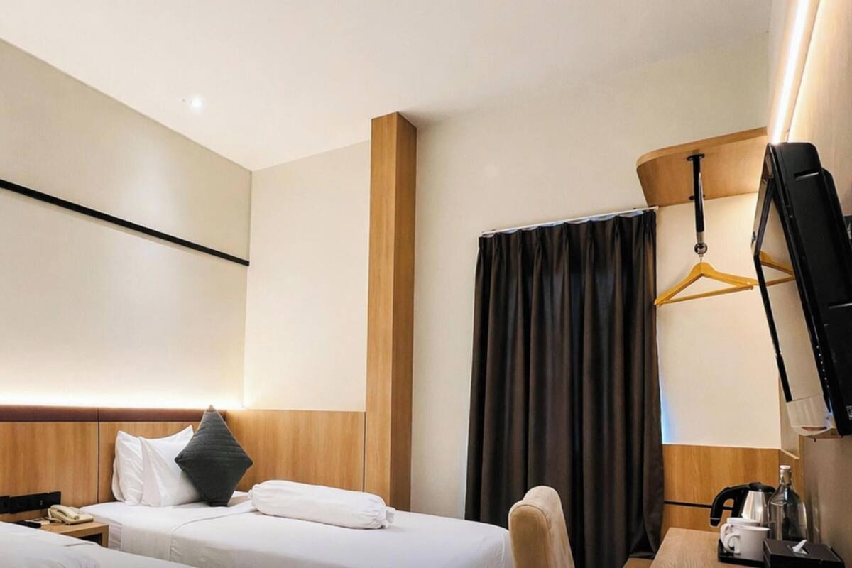 Une chambre d’hôtel avec deux lits, une tête de lit en bois, un téléviseur à écran plat fixé au mur, un bureau avec une chaise et un porte-manteau mural.