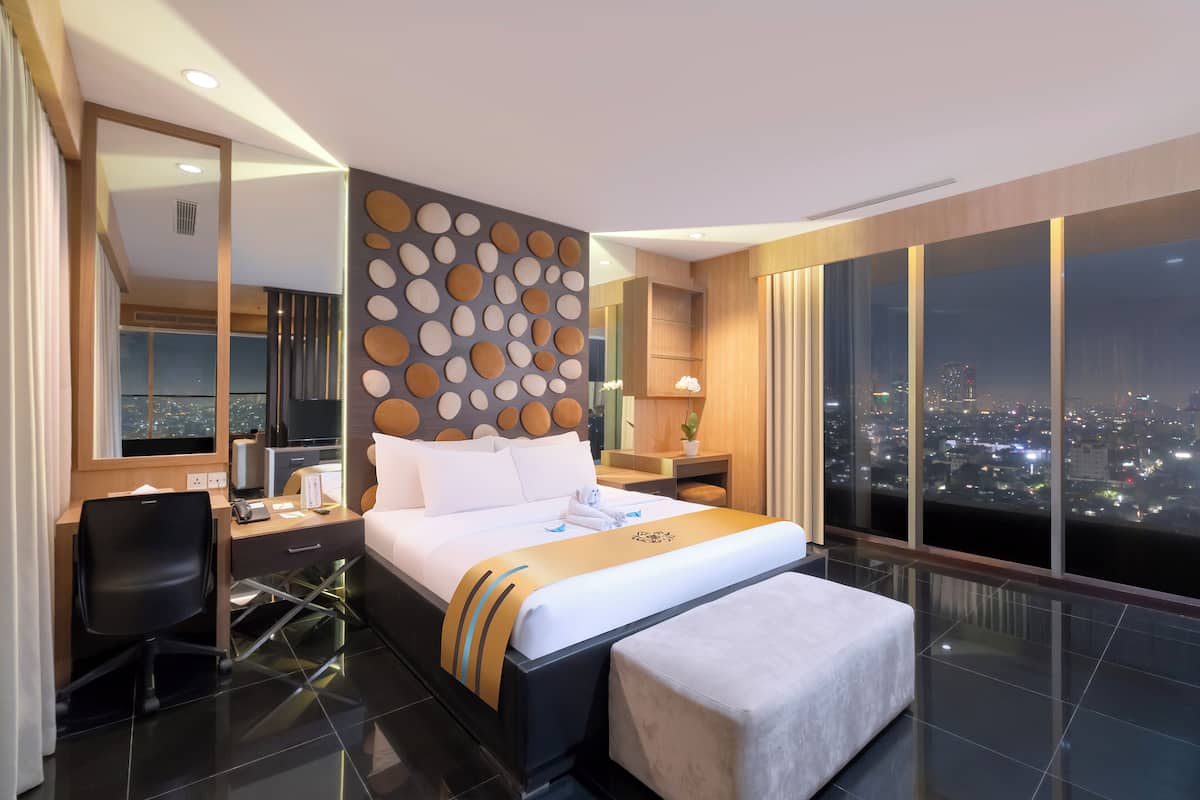 A modern hotel room with a large bed, a desk, a chair, and a city view through large windows.