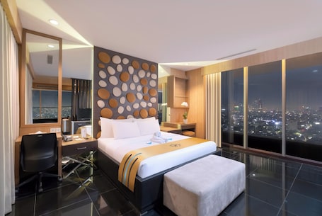 A modern hotel room with a large bed, a desk, a chair, and a city view through large windows.