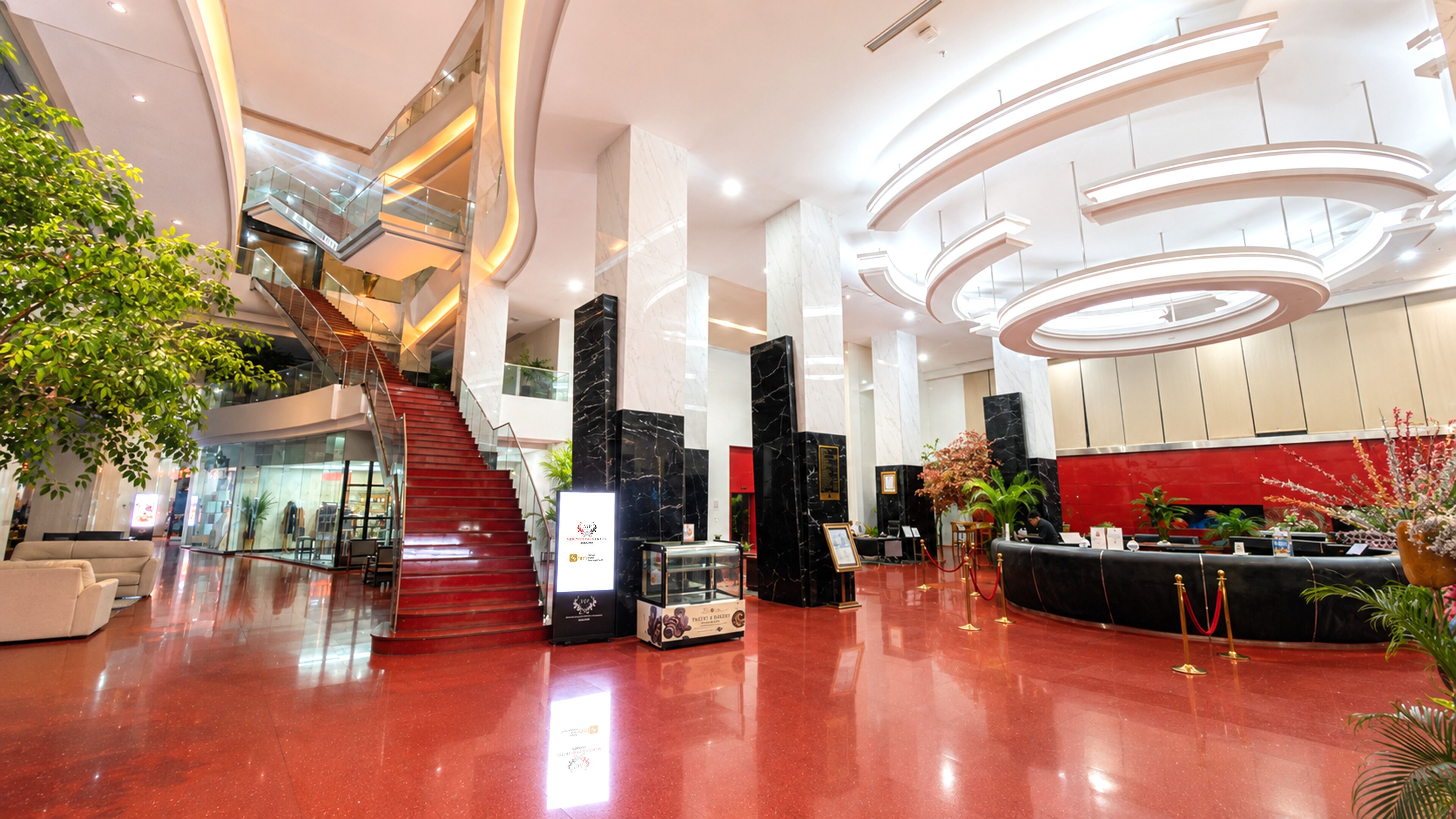 Un moderno vestíbulo de hotel con ascensor de vidrio, columnas de mármol, un mostrador de recepción y una zona con alfombra roja.