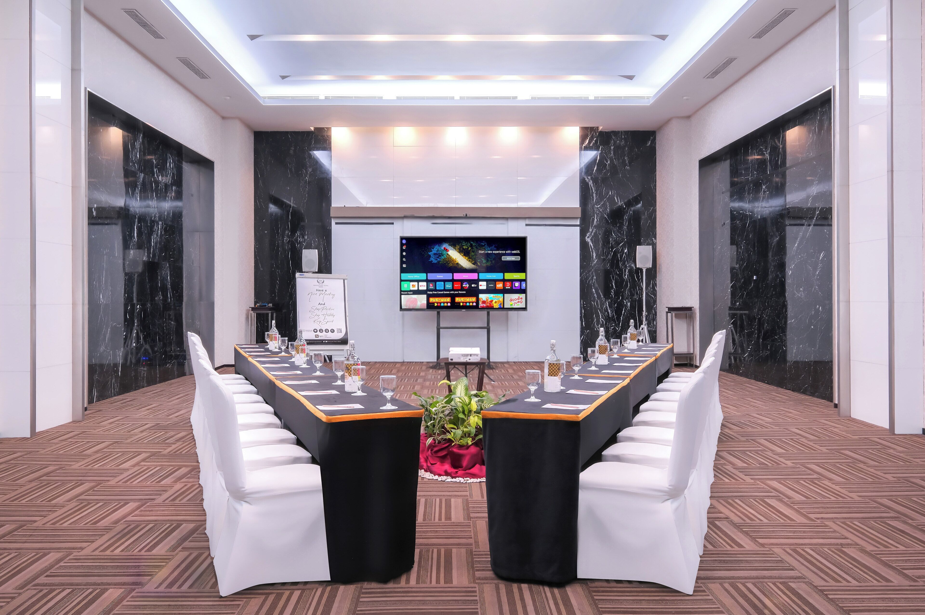 Un salón de conferencias con una larga mesa, sillas blancas, una pantalla y un letrero.