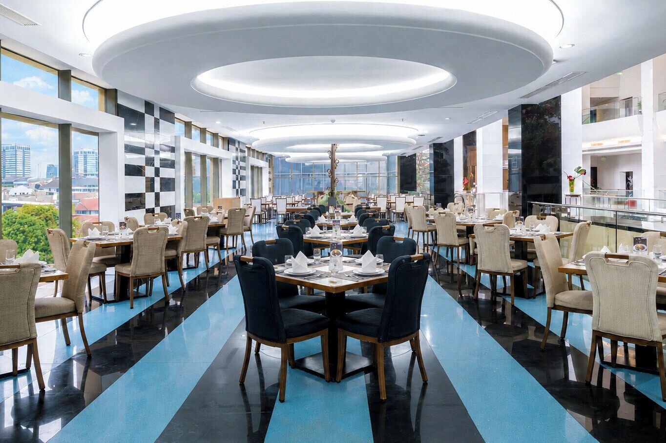 Un comedor amplio con varias mesas y sillas, ventanas grandes que ofrecen vistas a la ciudad y un diseño de techo moderno.
