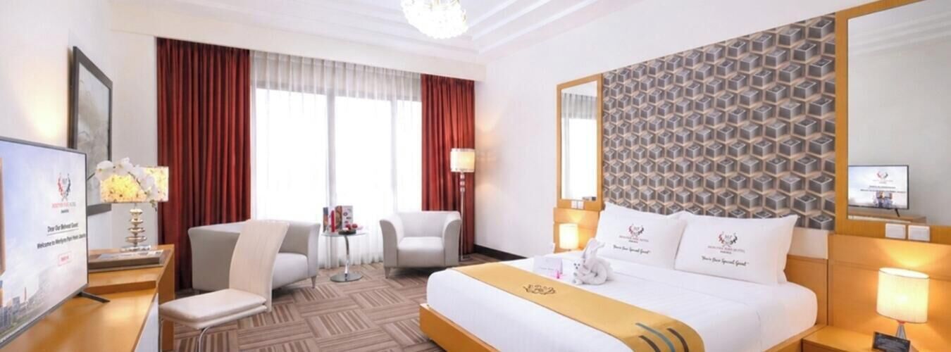 Una habitación de hotel con cama, dos sillas, escritorio, televisor y ventana con cortinas.