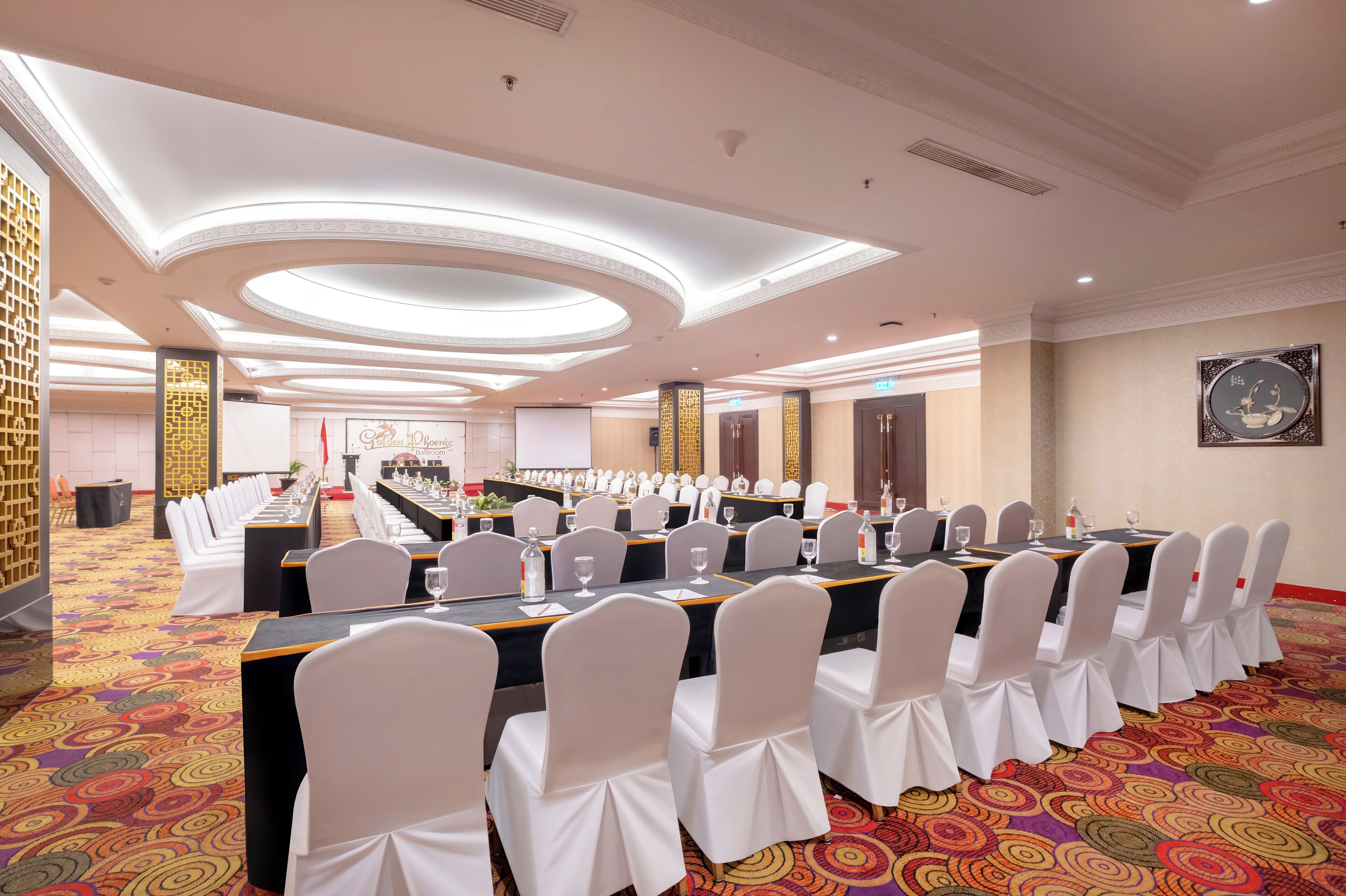 A conference room with long tables and white chairs, a patterned carpet, and a decorative ceiling.