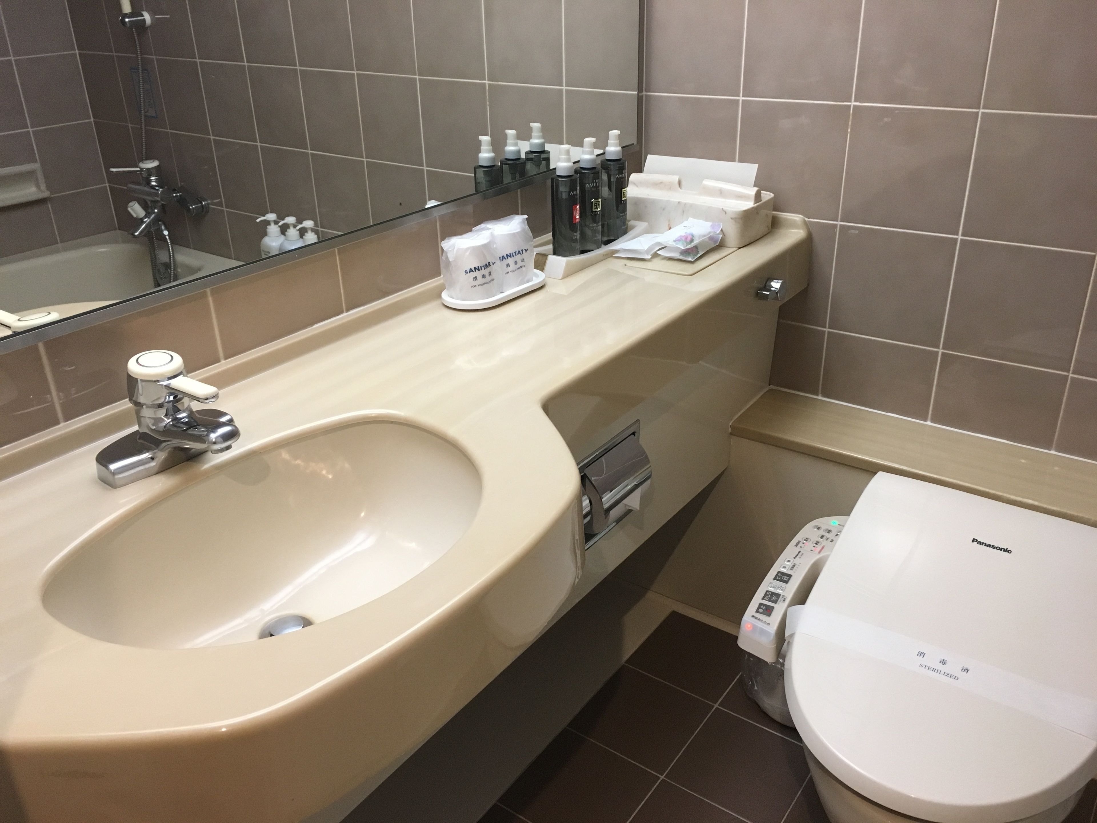 A modern bathroom with a large sink, a toilet, and a mirror. There are toiletries on the counter and a paper towel dispenser.