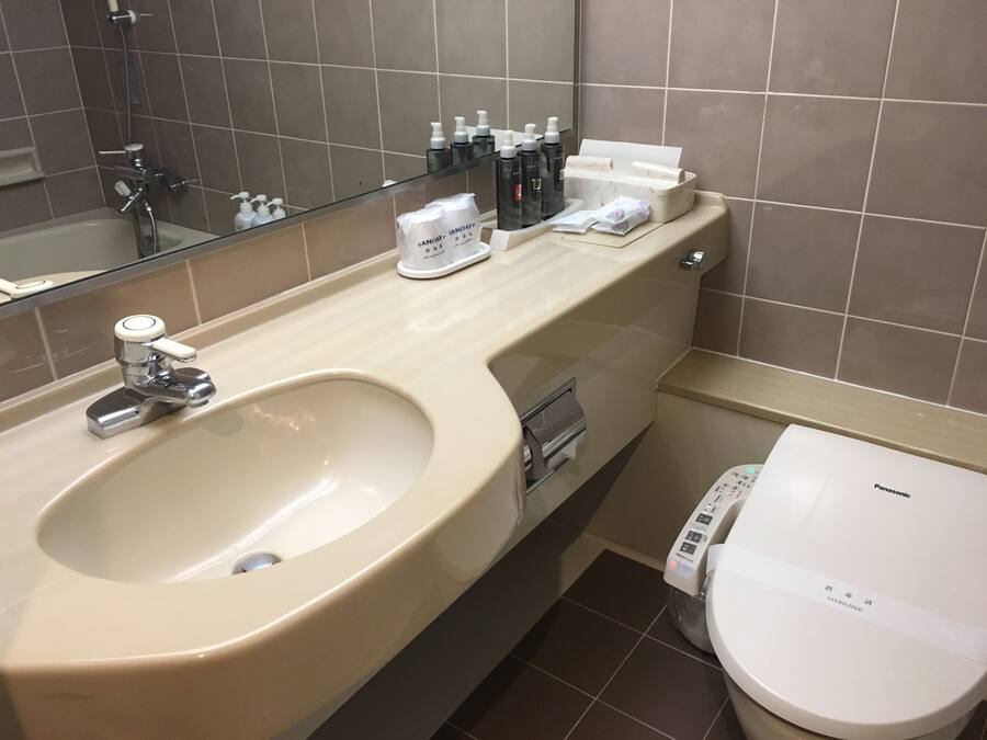 A modern bathroom with a large sink, a toilet, and a mirror. There are toiletries on the counter and a paper towel dispenser.