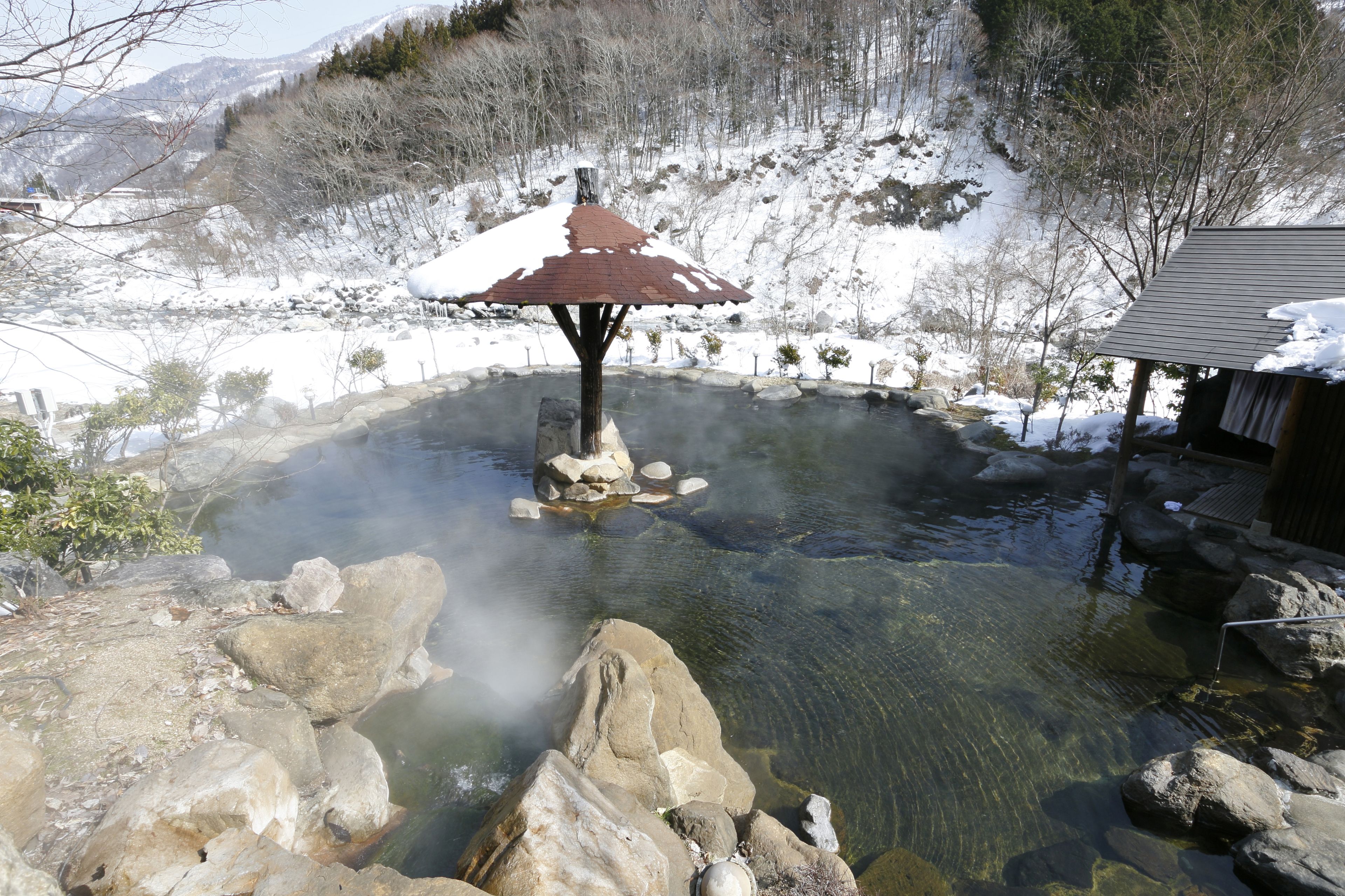 Un área de aguas termales con una estructura de piedra en el centro, rodeada de rocas y terreno cubierto de nieve.