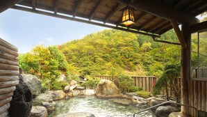 Un onsen japonés tradicional con estructura de madera, un manantial termal de piedra y vistas a una ladera boscosa.