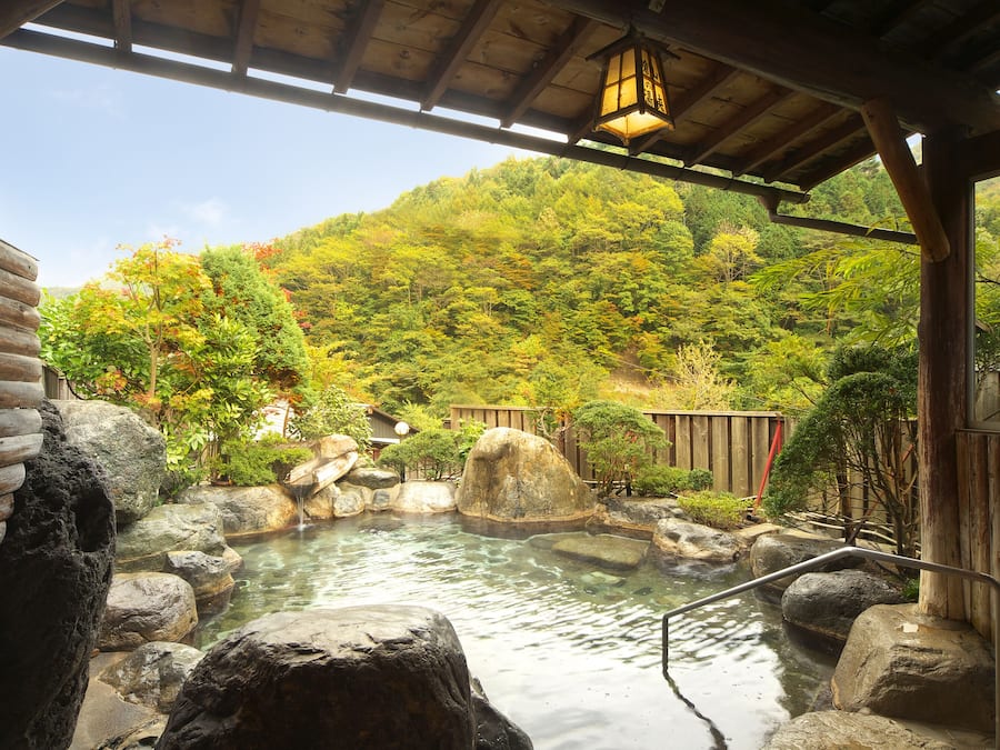 A traditional Japanese onsen with a wooden structure, a stone hot spring, and a view of a forested hillside.