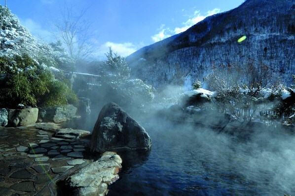 A serene hot spring area with a stone pathway, large rocks, and a mountain backdrop.