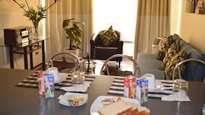 Un coin repas avec une table dressée pour deux personnes, comprenant un ordinateur portable, des assiettes de nourriture et des boissons. On y trouve une chaise avec un coussin à motifs et un canapé agrémenté d’un plaid décoratif.