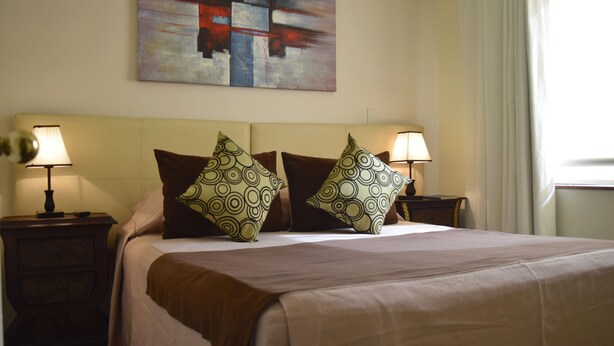 Un lit bien fait, recouvert d’une couvre-lit beige et agrémenté de deux coussins à motifs, avec deux tables de chevet de chaque côté, sur lesquelles sont posées des lampes.