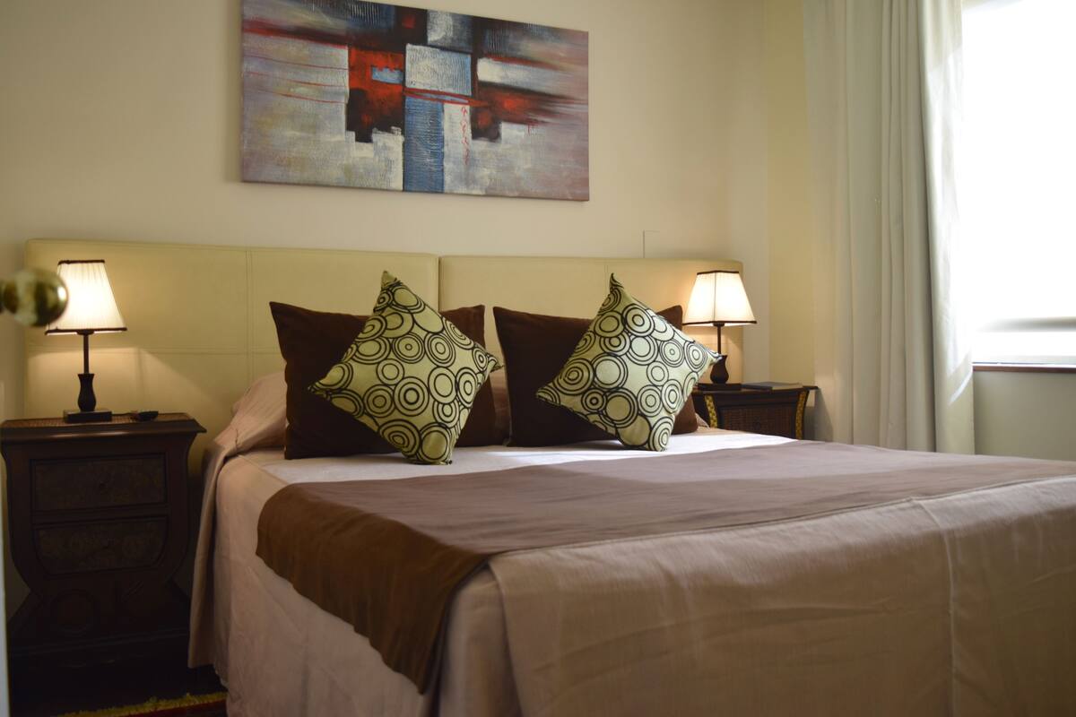 A neatly made bed with a beige bedspread and two patterned pillows, flanked by nightstands with lamps.